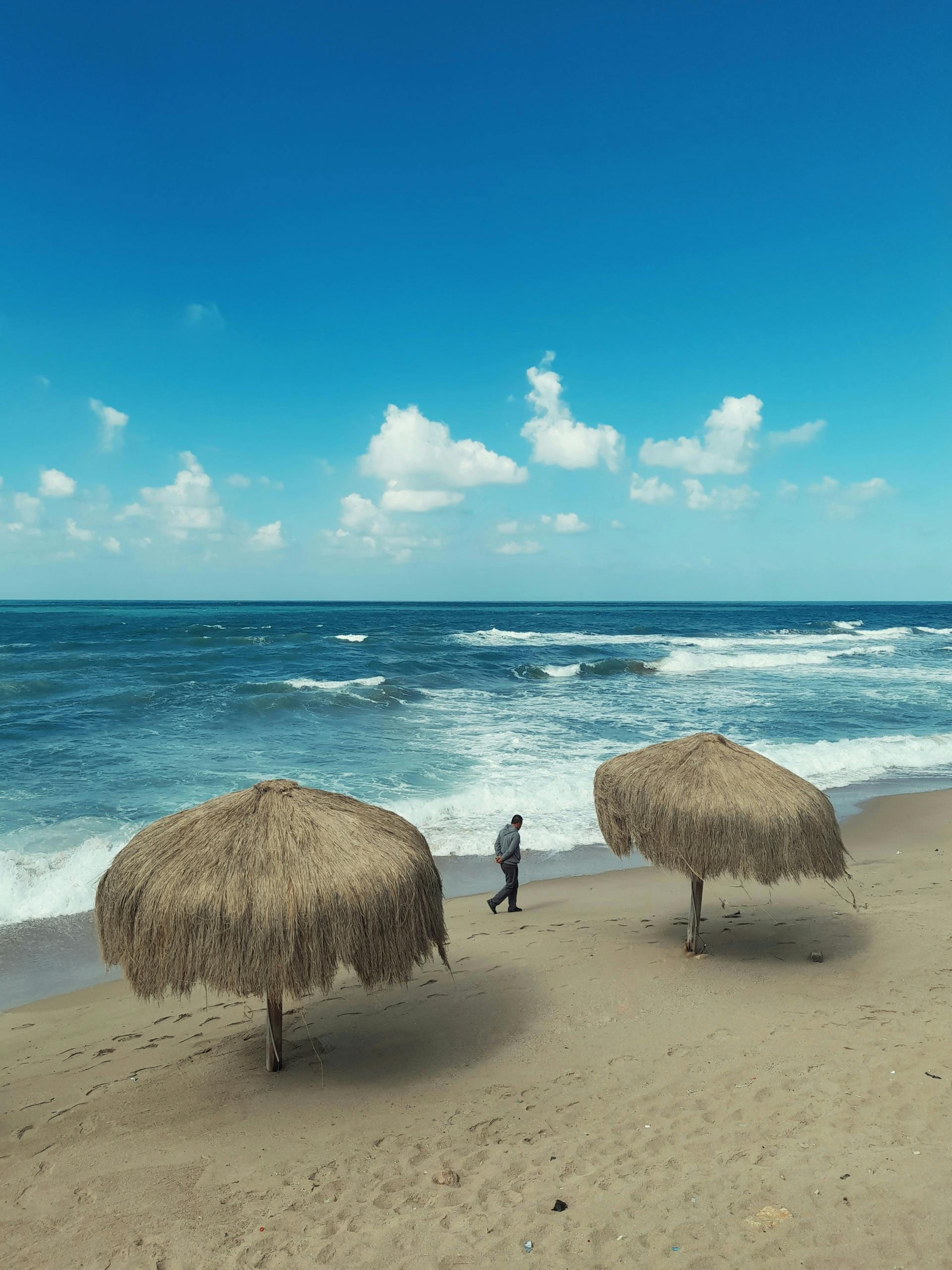 Man walking along Alexandria's coast with waves and thatched umbrellas under a clear sky.