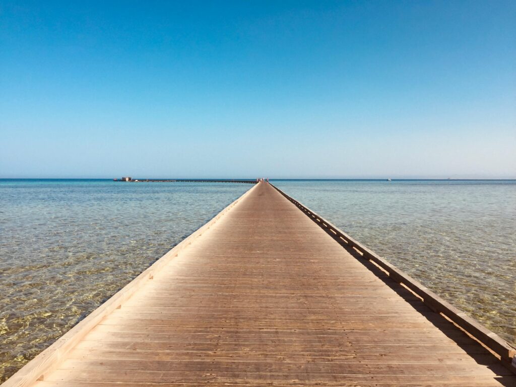 A wooden jetty extends into the crystal clear Red Sea, under a bright blue sky.