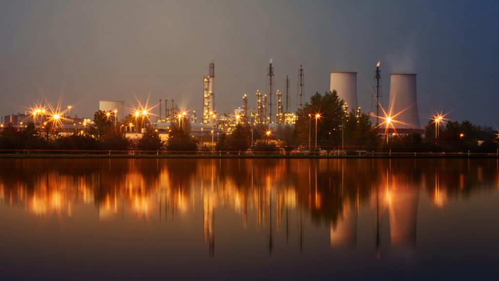 Industrial plant with reflection in water at twilight, showing smoke stacks and lights.