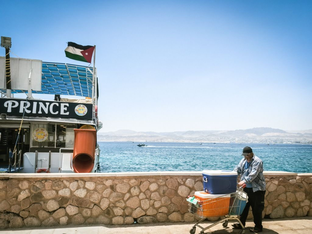 A man with a cart near a boat docked at the Red Sea in Aqaba, with scenic mountains in the background.