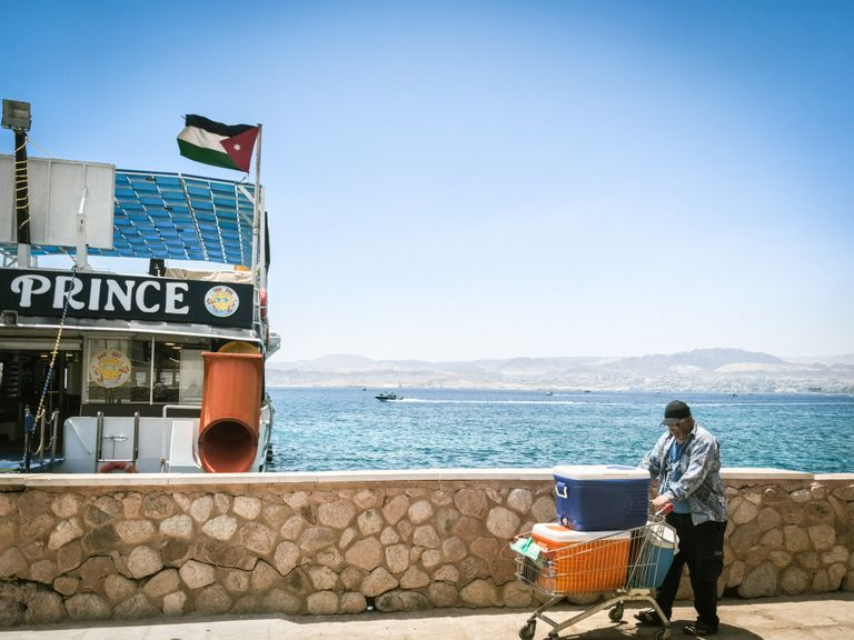 A man with a cart near a boat docked at the Red Sea in Aqaba, with scenic mountains in the background.