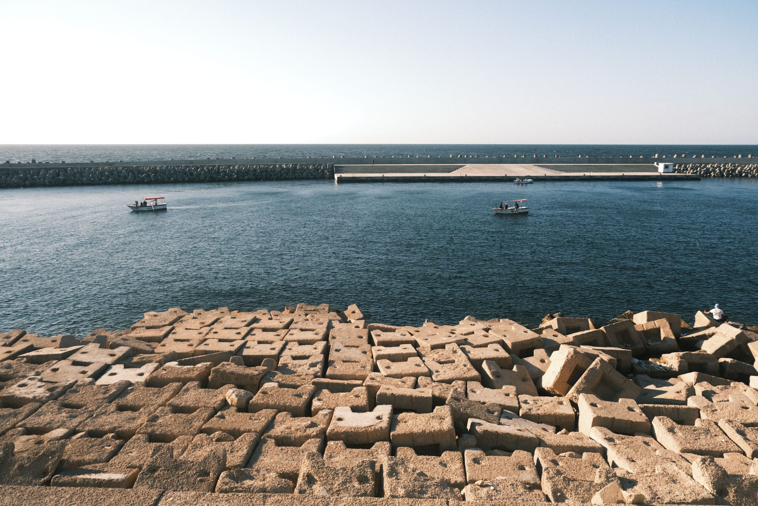 Biodiversity and Coastal Ecosystems Assessment for the Climate-Resilient ICZM Plan of Egypt's North Coast Tranquil scene of boats near a stone breakwater in Alexandria harbor under a clear sky.